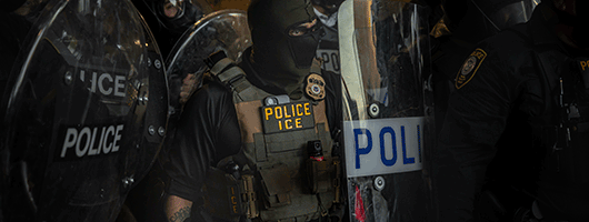 Photo of a masked ICE agent with a shield amid a larger group of officers with shields